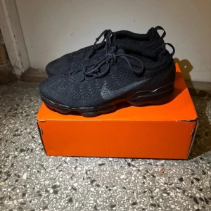 Nike Air Vapormax Flyknit svarta sneakers - Svarta Nike Air Vapormax Flyknit sneakers med stickad ovandel och synlig luftdämpning i sulan. Skorna har snörning och en sportig, modern look. Perfekta för dig som gillar streetwear och vill ha bekväma skor med cool design.