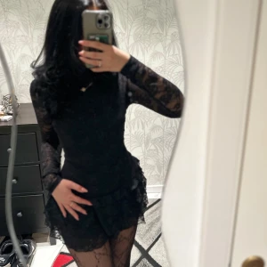 Svart spetsklänning med volanger - Säljer en svart kort klänning med långa spetsärmar och volangdetaljer. Klänningen har figurnära passform och flera lager volanger i kjolen. Har även ”inbyggda” shorts.