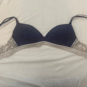 Mörkblå spets-bh från Creem - Säljer en mörkblå bh från Creem med tunna axelband och vackra spetsdetaljer i beige och blommigt mönster på sidorna. Bh:n har en mjuk och bekväm passform, perfekt för dig som gillar en feminin stil.