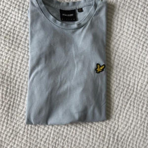 Ljusblå t-shirt från Lyle & Scott - Snygg ljusblå t-shirt från Lyle & Scott med klassisk rund hals och broderad gul logga på bröstet. Perfekt basic-plagg med stilren design som passar till det mesta. Bara att höra av sig vid minsta fundering!
