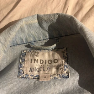 Ljusblå jeansjacka från Indigo Collection - Säljer en klassisk ljusblå jeansjacka från Indigo Collection. Jackan har knappar framtill, två bröstfickor med lock och knapp samt en snygg, tidlös design. Perfekt att slänga över en t-shirt för en avslappnad look.