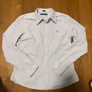 Ljusblå skjorta från Ralph Lauren - Säljer en stilren ljusblå skjorta från Ralph Lauren i skinny fit. Skjortan har långa ärmar och klassisk krage med knappar framtill. Den är dekorerad med det ikoniska broderade logotypen på bröstet. Perfekt för en elegant look.