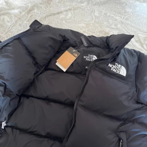 Svart pufferjacka från The North Face - Säljer en svart pufferjacka från The North Face med dragkedja och logga på bröstet. Jackan har en klassisk design med långa ärmar. Den är använd ett par gånger men har satt tillbaka taggen ändå och har gjort jackan precis som helt ny. Det är bara att höra av sig kring frågor och funderingar! Pris går också att diskutera