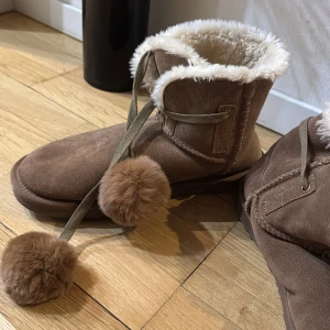 Bruna boots med pälsdetaljer - Säljer ett par mysiga bruna boots med fluffig päls på insidan och dekorativa bollar. Perfekta för kalla dagar och ger en stilfull touch till din outfit. Använda endast 1gång så i princip som nya. Ord pris 1200kr
