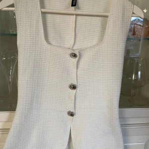 Vit topp med silvriga knappar - Snygg vit topp från H&M med tre dekorativa knappar framtill. Västen har en elegant och stilren design, perfekt för att lyfta vilken outfit som helst. Helt ny men saknar lapp, säljer då den var för stor🤍