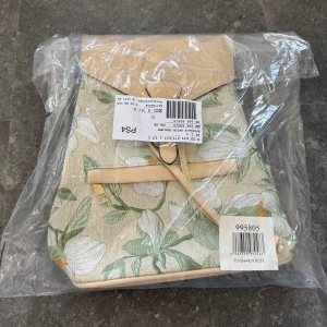 Blommig ryggsäck med beige detaljer - Snygg ryggsäck med blommigt mönster i grönt och vitt. Väskan har beige detaljer och justerbara axelremmar. Perfekt för en stilren look. Helt oanvänd