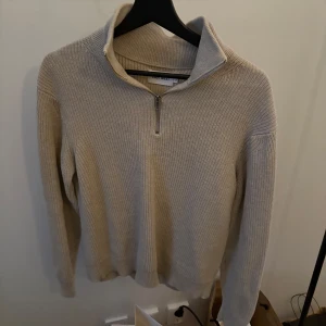 Beige stickad tröja med dragkedja - Säljer en stilren beige stickad tröja från East west. Tröjan har en dragkedja vid halsen och är tillverkad i 100% bomull. Perfekt för kyligare dagar med sin långa ärmar och mjuka material. Den är strl M men skulle mer säga S.