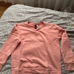 Rosa sweatshirt från Adidas - Säljer en snygg rosa sweatshirt från Adidas med deras logga och texten 'Athletics' på framsidan. Tröjan har långa ärmar och en rund halsringning. Perfekt för en avslappnad stil.