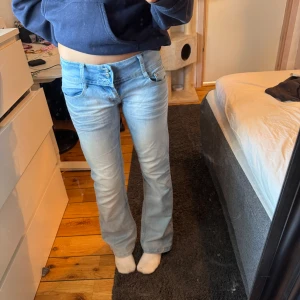 Ljusblå low waist jeans - Snygga ljusblå, low waist, bootcut jeans med unika blomformade knappar. Byxorna har en klassisk femficksdesign och en bekväm passform. Perfekta för en avslappnad stil. Innerbenslängden: ca 81cm. Midjemått: ca 38cm. Knapp saknas längst ner, men tycker inte det är särskilt synligt, därav lägre pris. 