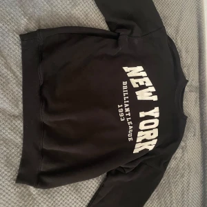 Svart sweatshirt från Bershka - Säljer en svart sweatshirt från Bershka med texten 'New York Brilliant League 1993' i vitt på framsidan. Tröjan har långa ärmar och ribbade muddar vid ärmslut och nederkant. Perfekt för en avslappnad stil.