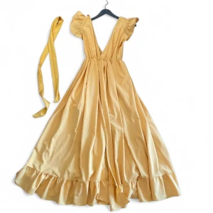 Gul långklänning med volangdetaljer - Vacker gul långklänning med volangdetaljer och djup v-ringning. Klänningen har en matchande knytband och är perfekt för somriga tillfällen. Den är ärmlös och har en luftig passform som ger en romantisk känsla. Passar perfekt till balen! Helt ny utan prislapp.  One-size i storlek men skulle säga att den är som en S. 