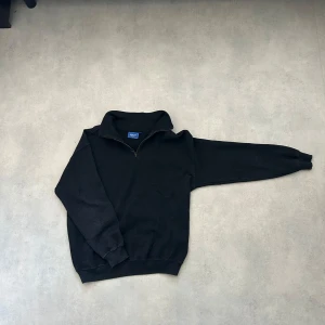 Svart tröja med dragkedja från Gildan - Säljer en svart tröja från Gildan med halv dragkedja och hög krage. Perfekt för kyliga dagar med sin ribbade struktur och långa ärmar. Enkelt och stilrent plagg för garderoben.