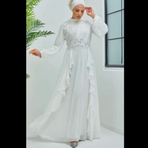 Vit långklänning med spetsdetaljer - Elegant vit långklänning med vackra spetsdetaljer och volanger. Klänningen har långa ärmar och en flytande design som ger en romantisk känsla. Perfekt för speciella tillfällen.