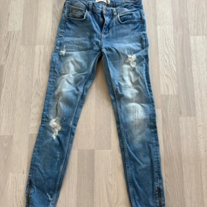 Blå jeans från Gina Tricot - Säljer ett par blå jeans från Gina Tricot med slitna detaljer och dragkedja vid anklarna. Modellen heter Kristen och har en snygg, modern look. Perfekta för en avslappnad stil. Använda men fortfarande bra skick. Storlek 28/30 Skinny jeans
