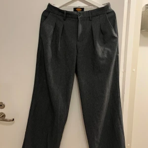 Grå randiga kostymbyxor från Urban Outfitters - Snygga grå kostymbyxor från Urban Outfitters med diskret randigt mönster. Byxorna har en klassisk passform med mid waist och raka ben. Perfekta för en stilren look.