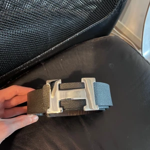 Svart bälte med silverfärgat spänne - Snyggt svart bälte med ett stort, silverfärgat H-format spänne. Perfekt för att ge en stilren touch till din outfit. Bältet är tillverkat i skinn och har en klassisk design som passar till många olika stilar.