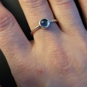 Silverring med blå sten - Elegant silverring med en vacker blå sten i centrum. Ringen har en stilren design med en slät yta och en rund infattning för stenen. Perfekt för att ge en subtil färgklick till din outfit.