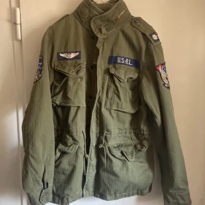 Grön militärinspirerad jacka från Ralph Lauren - Säljer en grön militärinspirerad jacka från Ralph Lauren med flera broderade märken och detaljer. Jackan har en hög krage och knäppning framtill. Perfekt för en cool och avslappnad stil. 10/10 skick. 
