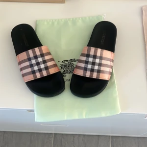 Rutig sandaler från Burberry - Snygga sandaler från Burberry med klassiskt rutigt mönster i beige, svart och rött. Perfekta för en avslappnad stil med en touch av lyx. Sandalerna har en bred rem över foten och en bekväm svart sula.