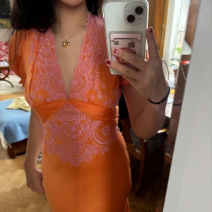Orange klänning med spetsdetaljer - Säljer en snygg orange klänning med v-ringad design och vackra spetsdetaljer i lila. Klänningen har korta ärmar och en öppen rygg med knytband. Storlek 38