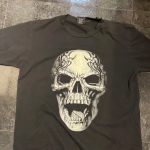 Svart t-shirt med dödskallemotiv - Cool svart t-shirt med ett stort, detaljerat dödskallemotiv på framsidan. Perfekt för en edgy look. T-shirten har korta ärmar och en klassisk rund halsringning.
