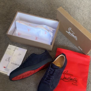 Mörkblå sneakers från Christian Louboutin - Snygga mörkblå sneakers från Christian Louboutin med den ikoniska röda sulan. Skorna har snörning och är tillverkade i mocka, vilket ger en lyxig känsla. Perfekta för att ge din outfit en stilfull touch.