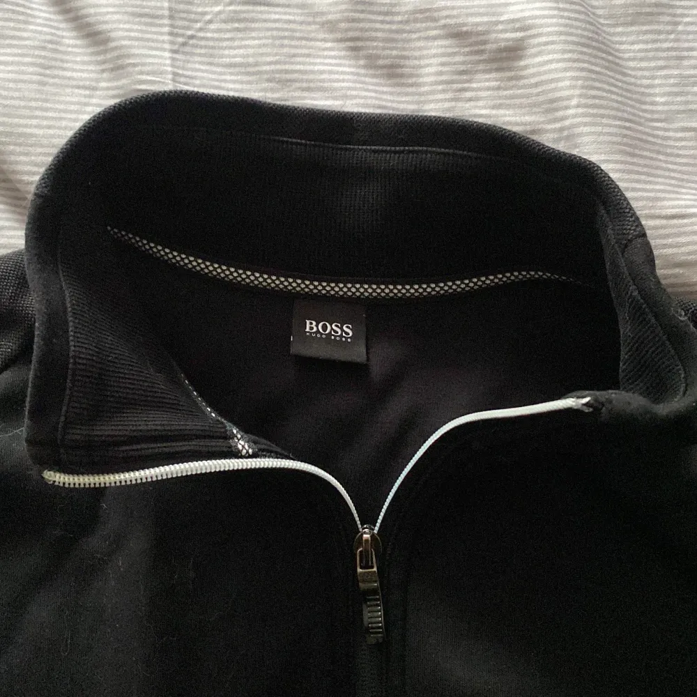 Snygg svart jacka från Boss med dragkedja framtill och hög krage. Perfekt för en stilren look. Jackan har långa ärmar och två fickor på sidorna. Stryker tröjan efter försäljning.. Neuletakit & Villapaidat.