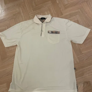 Vit kortärmad pikétröja från Burberry - Säljer en stilren vit pikétröja från Burberry med klassiskt rutigt mönster på kragen och bröstfickan. 