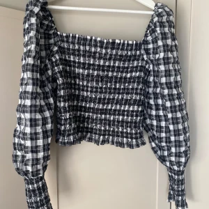 Svartvit rutig offshoulder topp från H&M - Säljer en snygg svartvit rutig offshoulder topp från H&M. Toppen har långa ärmar och en smockad design som ger en tight passform. Perfekt för en trendig look!