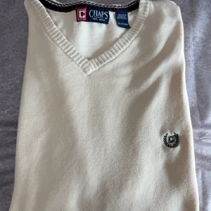 Beige ärmlös tröja från Chaps - Säljer en stilren beige ärmlös tröja från Chaps med v-ringning och broderad logga på bröstet. Perfekt för lager-på-lager-stil eller sommarens varma dagar. Tillverkad i mjuk bomull för extra komfort. Passar även XL