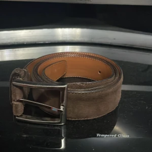 Brunt bälte med silverfärgat spänne - Snyggt brunt bälte i mocka med ett stilrent silverfärgat spänne. Perfekt för att ge en elegant touch till din outfit. Bältet har en klassisk design och passar till många olika stilar.