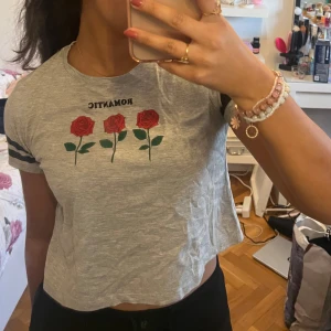 Grå t-shirt med rosor och text - Snygg grå t-shirt från Pull&Bear med tryck av tre röda rosor och texten 'ROMANTIC' på bröstet. T-shirten har korta ärmar med svarta ränder och en croppad passform. Perfekt för en avslappnad stil!