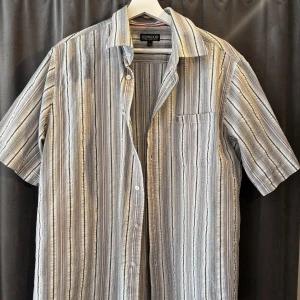 Randig kortärmad skjorta från Redwood - Säljer en stilren randig skjorta från Redwood i grå och beige toner. Skjortan har korta ärmar och en klassisk krage med knappar framtill. Perfekt för en avslappnad stil.