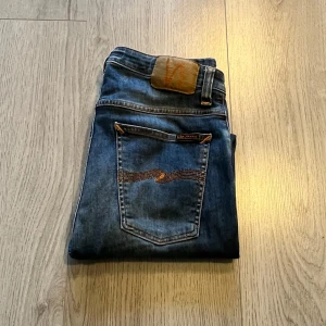 Blå jeans från Nudie Jeans - Snygga blå jeans från Nudie Jeans med klassisk femficksdesign och orange sömmar. Jeansen har en straight passform och är tillverkade i slitstarkt denim. Perfekta för en avslappnad stil.