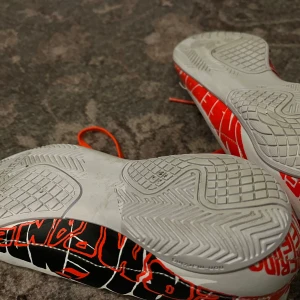 Vita och röda fotbollsskor från Adidas - Snygga fotbollsskor från Adidas i vitt med röda och svarta detaljer. Skorna har en iögonfallande design med text på sulan och röda snören. Perfekta för inomhus (fotsall osv)  med en stilren look.