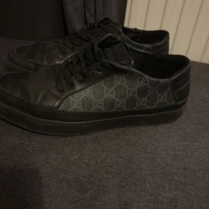 Gucci skor - Snygga svarta sneakers från Gucci med ett diskret monogrammönster på sidan. Skorna har en klassisk snörning och är tillverkade i en kombination av skinn och textil. Perfekta för en stilren look.