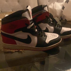 Nike Air Jordans i rött, svart och vitt - Snygga Nike Air Jordans i klassisk röd, svart och vit färgkombination. Skorna har en hög design med snörning och den ikoniska Air Jordan-loggan på sidan. Perfekta för dig som vill ha en stilren och sportig look.