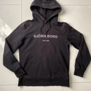 Svart hoodie från Björn Borg - Säljer en stilren svart hoodie från Björn Borg med tryck på framsidan. Tröjan har en klassisk design med huva och dragsko samt en stor ficka framtill. Perfekt för en avslappnad look. Storlek M och använt sällan så är i bra skick!