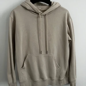 Beige hoodie med ficka - Säljer en stilren beige hoodie med huva och dragsnören. Tröjan har en stor magficka och ribbade muddar vid ärmslut och nederkant. Perfekt för en avslappnad look. 