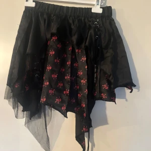 Kjol med små döskallar i onesize - Säljer en svart kortkjol med asymmetrisk och trasig look. Kjolen har röda dödskallar som mönster och snörningsdetaljer på sidorna. Resår i midjan och flera lager tyg ger en cool och edgy stil.
