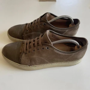 Ljusbruna sneakers i mocka från Lanvin - Säljer ett par ljusbruna sneakers från Lanvin i mocka med snörning och rund tå. Skorna har en beige sula och detaljer i skinn på tån. Perfekta för dig som gillar stilrena och klassiska sneakers.