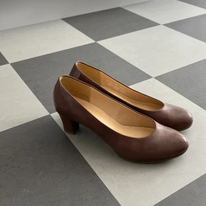 Bruna pumps från Gabor - Säljer ett par klassiska bruna pumps från Gabor med rund tå och klack. Skorna är i skinn och har en stilren design som passar till många olika outfits. Perfekta för dig som gillar en tidlös och elegant look.