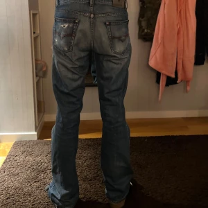 Blå jeansbyxor från Rodrigues  - Säljer ett par klassiska blå jeansbyxor från Rodrigues med raka ben och normal passform. Jeansen har slitningar och hål på framsidan samt detaljerade bakfickor. Perfekta för en avslappnad stil.