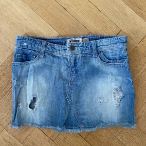 Blå jeanskjol från Stradivarius - Säljer en blå lågmidjad jeanskjol från Stradivarius med slitna detaljer och rå kant nertill. Kjolen har klassiska fem fickor och bälteshällor. Perfekt för dig som gillar en avslappnad och trendig stil.