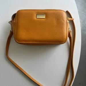 Gul axelväska med gulddetalj - Säljer en snygg gul axelväska med justerbar rem och två stora fack med dragkedja. Väskan har en guldfärgad detalj framtill och på spännena. 