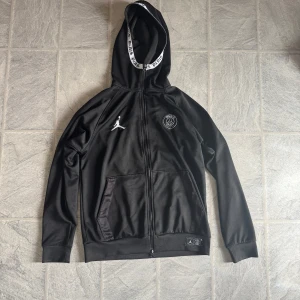 Svart PSG x Jordan Zip Up - Svart huvtröja med dragkedja från Jordan x PSG. Har vita loggor på bröstet, huva med PSG-tryck på kanten och två fickor framtill. Sportig look med ribbade muddar och mjukt material. Säljer denna på grund av att den är för liten.