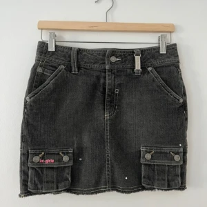 Svart jeanskjol - Cool svart jeanskjol med två stora fickor framtill och broderad text på baksidan. Kjolen har hällor för bälte, knappstängning och dekorativa sömmar. Plisserad nederdel baktill ger en extra detalj. 