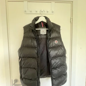 Svart dunväst från Moncler - Snygg svart dunväst från Moncler med hög krage och dragkedja framtill. Västen har Moncler-logga på bröstet och innerfoder med serietidningsmönster. Perfekt för lager-på-lager och stilren look.