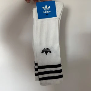Vita strumpor från Adidas med svarta ränder - Klassiska vita Adidas-strumpor med svarta ränder vid ankeln och Adidas-logga framtill. Ribbstickad design som sitter bra på foten. Perfekta för sport eller vardag.
