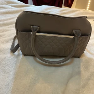 Beige quiltad handväska med kedjedetalj - Säljer en beige handväska med quiltad framsida och snygga silverfärgade kedjedetaljer på sidorna. Väskan har både handtag och en justerbar axelrem samt dragkedja upptill. Perfekt för dig som vill ha en stilren och rymlig accessoar.